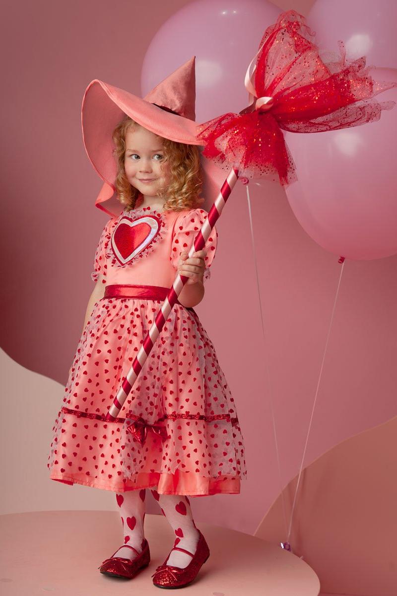 Child in a pink dress with heart design, holding a red and white wand against a pink background.