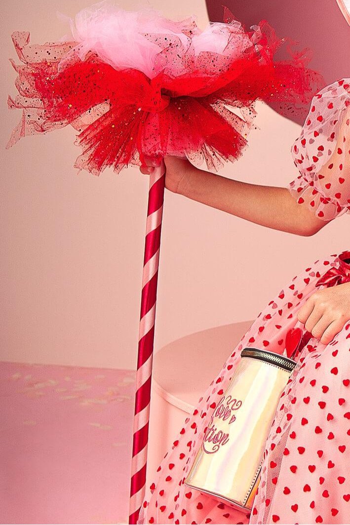 Child in a red and white polka dot dress holding a large red bow with a pink background